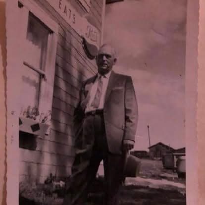 Sepia-toned archival photograph of a man in a suit standing in front of the historic 'EATS' diner storefront on the Watson's property